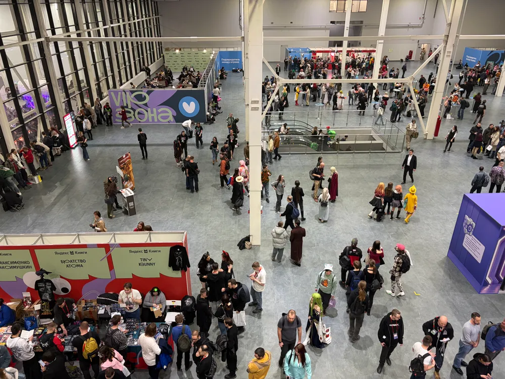 На картинке изображено большое количество людей, находящихся в помещении, возможно, на мероприятии или выставке. Видны стенды и различные активности.