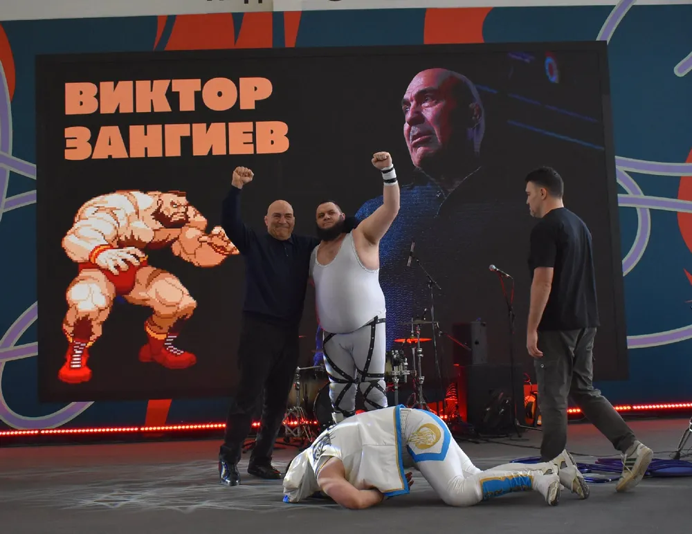 Изображение на сцене с людьми, один из них в костюме борца, другие участники в обычной одежде, фон с надписью \