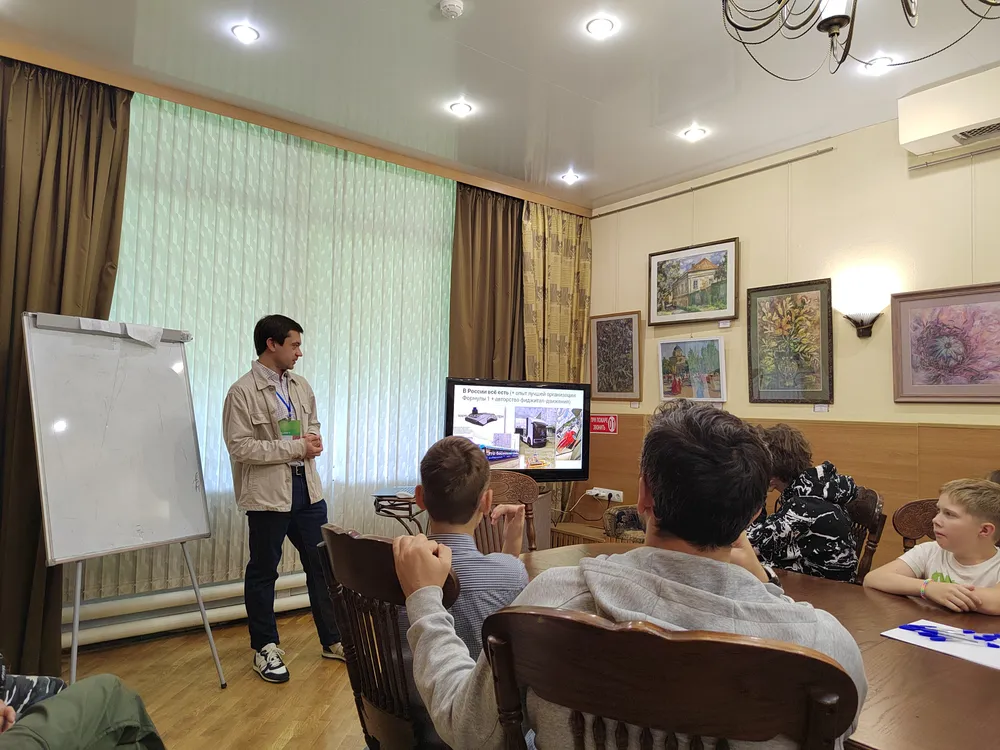 Мужчина, выступающий на презентации перед аудиторией. На стене картины. В комнате есть стол и стулья.