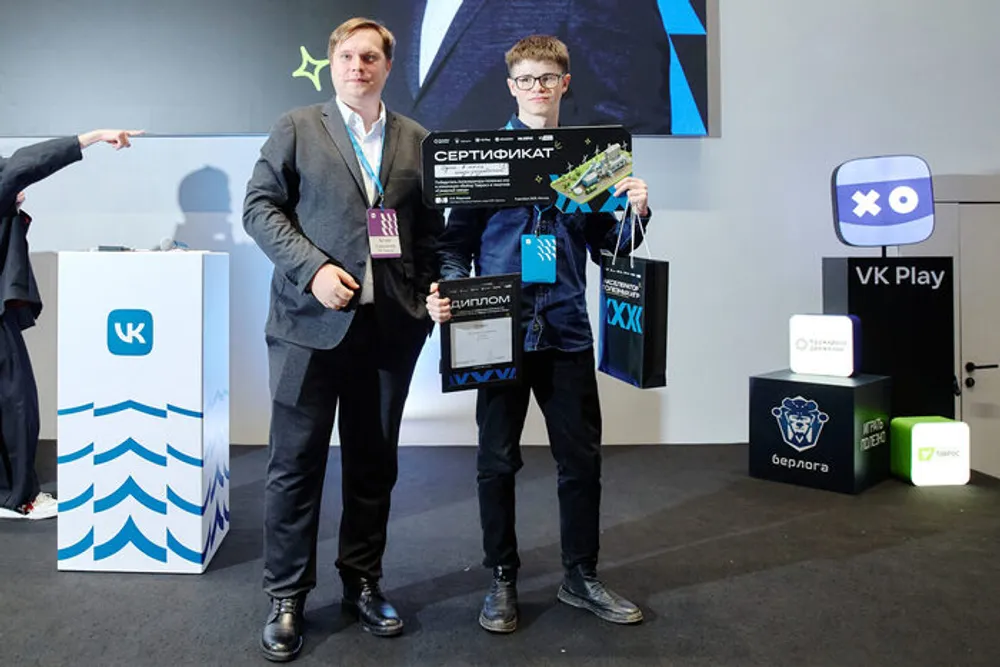 Два человека на мероприятии, один из них держит сертификат. Фон с логотипами и рекламой.