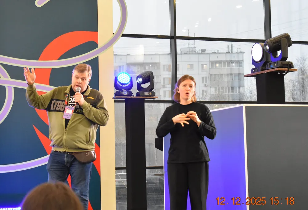 На изображении мужчина, говорящий на сцене, и женщина, выполняющая жестовый язык. Освещение и сцена с фоновыми элементами.