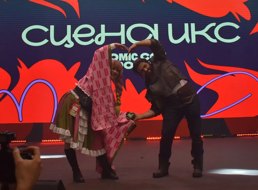 На изображении два человека, которые участвуют в выступлении на сцене. Они одеты в костюмы, фон содержит текст и графику, относящиеся к комиксам или мероприятиям поп-культуры.