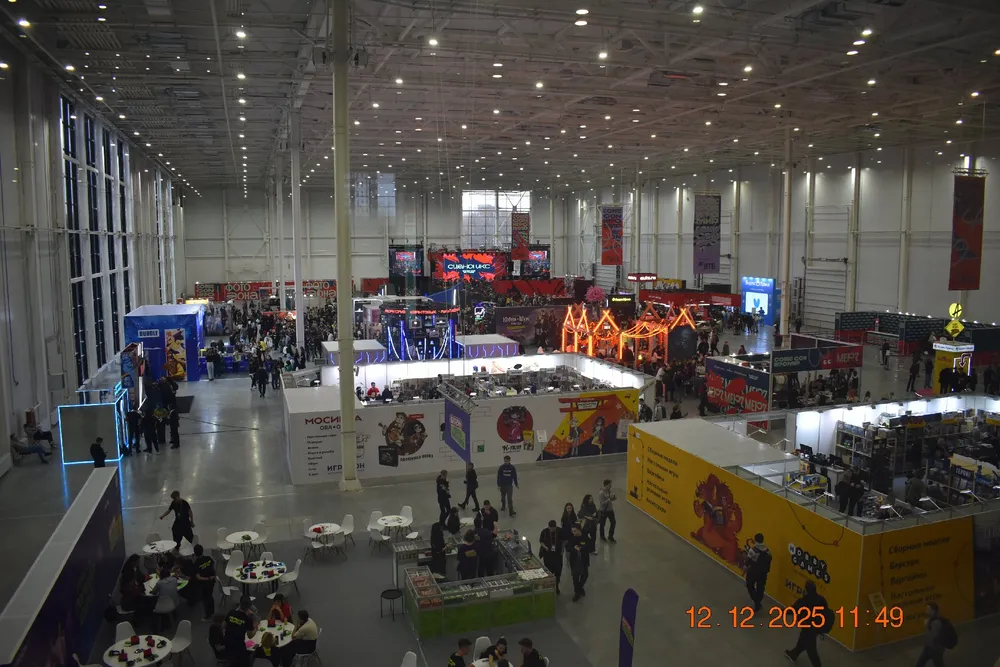 На картинке изображен большой зал с выставкой, в котором находятся стенды и посетители. Видны различные рекламные конструкции и много людей.