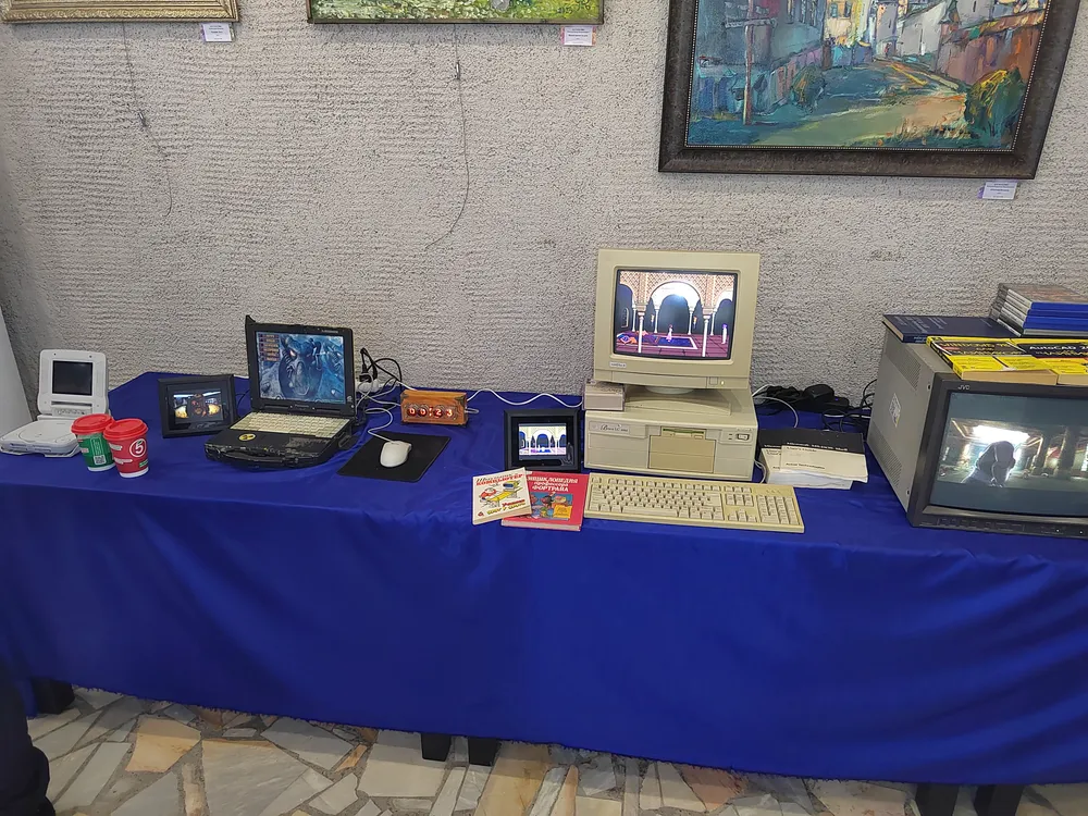 Выставка старых компьютеров и техники, включая монитор, клавиатуру, и другие устройства на столе с синим покрытием.