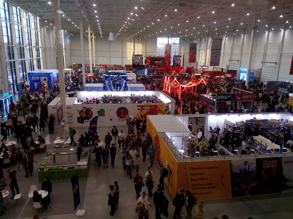 Изображение показывает большое количество людей на выставке или мероприятии в помещении, с различными стендами и освещением.