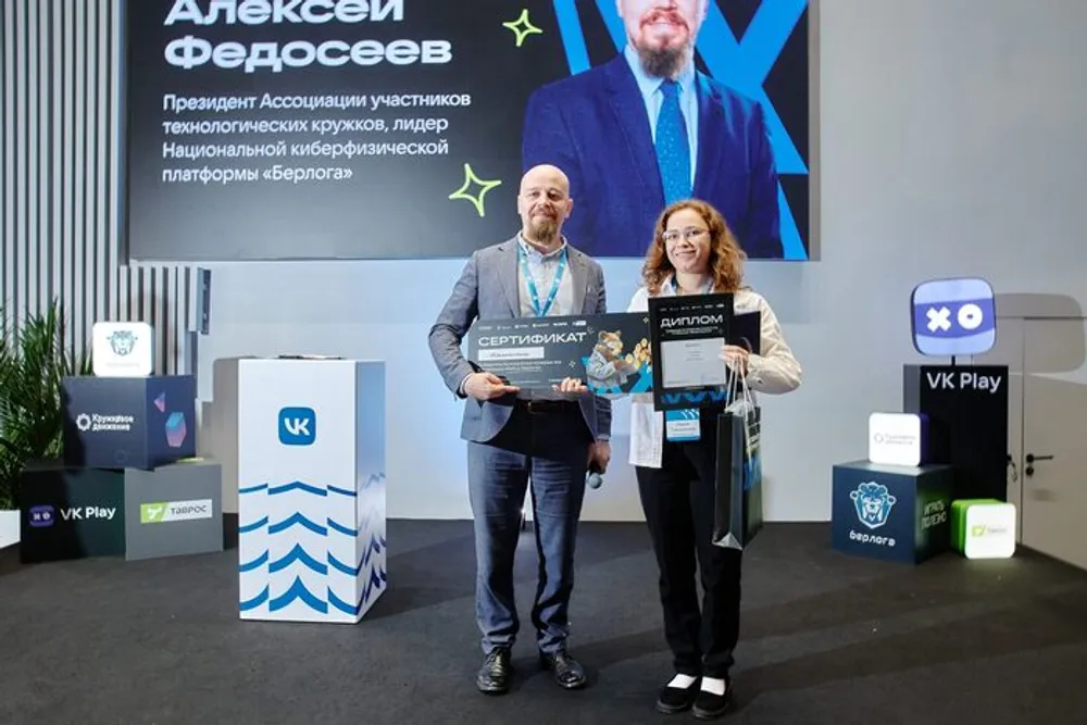 На изображении два человека на мероприятии, один из них держит табличку с текстом. Фон включает логотипы и название компании.