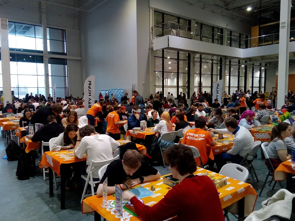 Изображение большого количества людей, сидящих за столами в помещении, вероятно, на мероприятии или фестивале. Люди занимаются различными активностями.