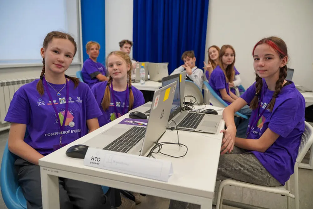 Группа подростков сидит за столами с ноутбуками, в фиолетовых футболках, в комнате с белыми стенами и синими занавесками.
