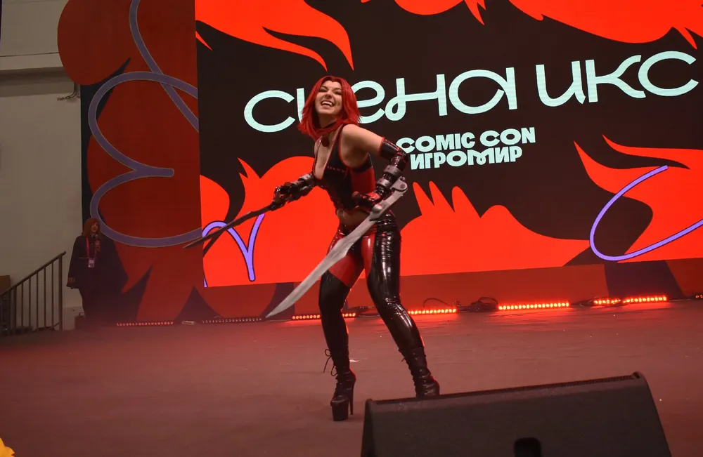 На изображении показана женщина в костюме, которая позирует на сцене. Фон содержит текст и элементы дизайна, указывающие на мероприятие, вероятно, связанное с комиксами или играми.