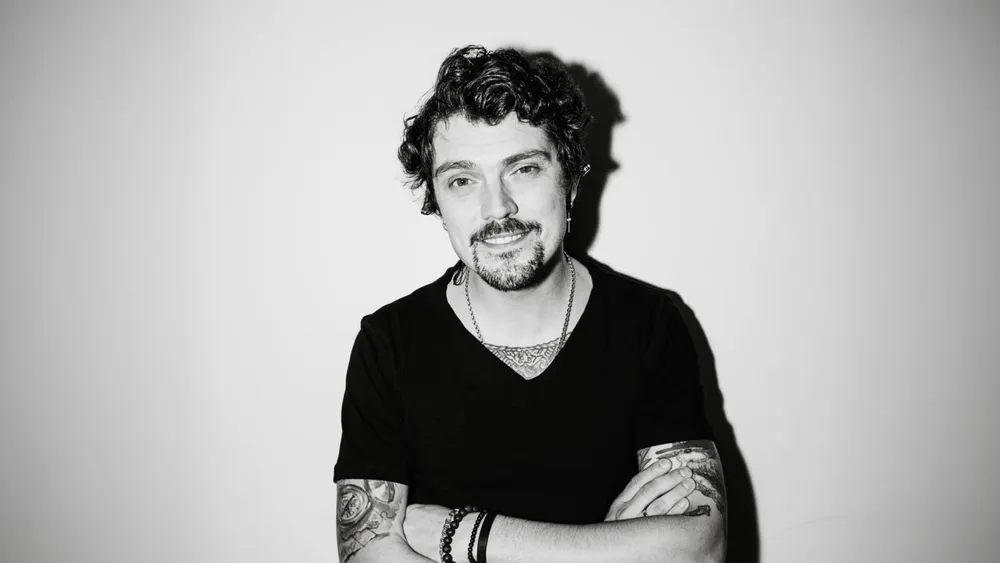 Человек с тёмными волосами и татуировками, одет в чёрную футболку, позирует с перекрещенными руками на фоне светлой стены.