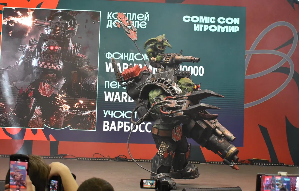 На изображении представлен человек в костюме персонажа из игры Warhammer 40,000 на мероприятии Comic Con. В фоне видно декорации и людей, снимающих на камеры.
