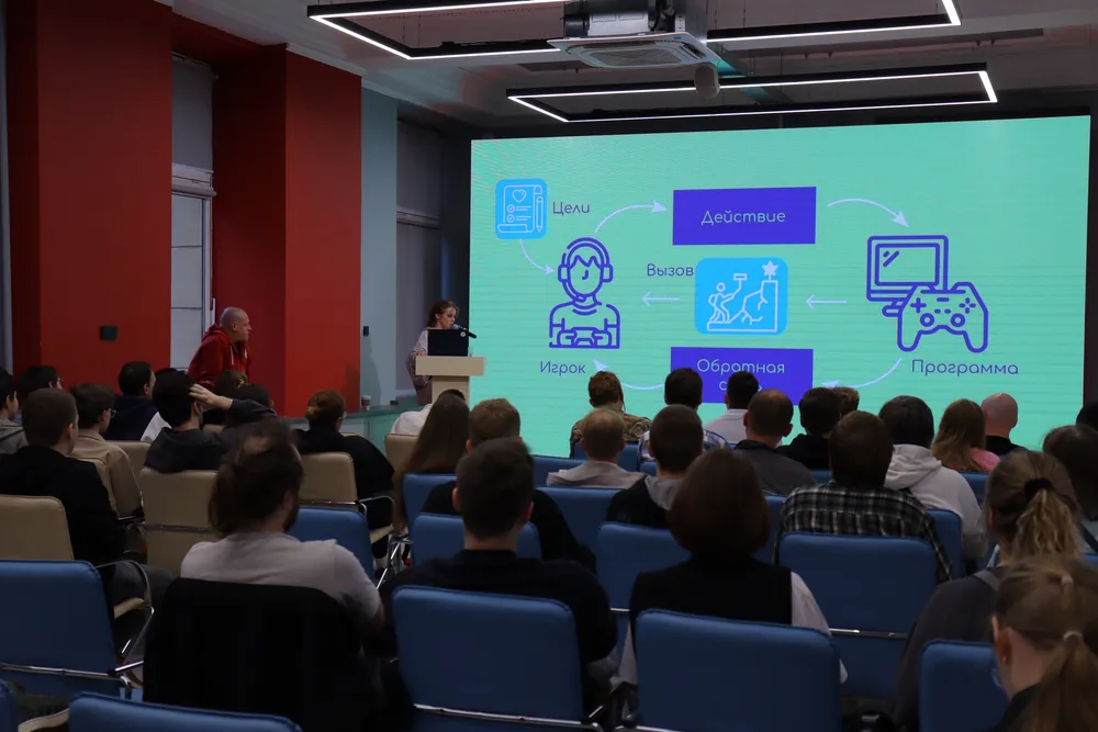 Изображение демонстрирует аудиторию на мероприятии, где выступает спикер. На заднем плане видно презентацию, возможно, о технологиях или играх.