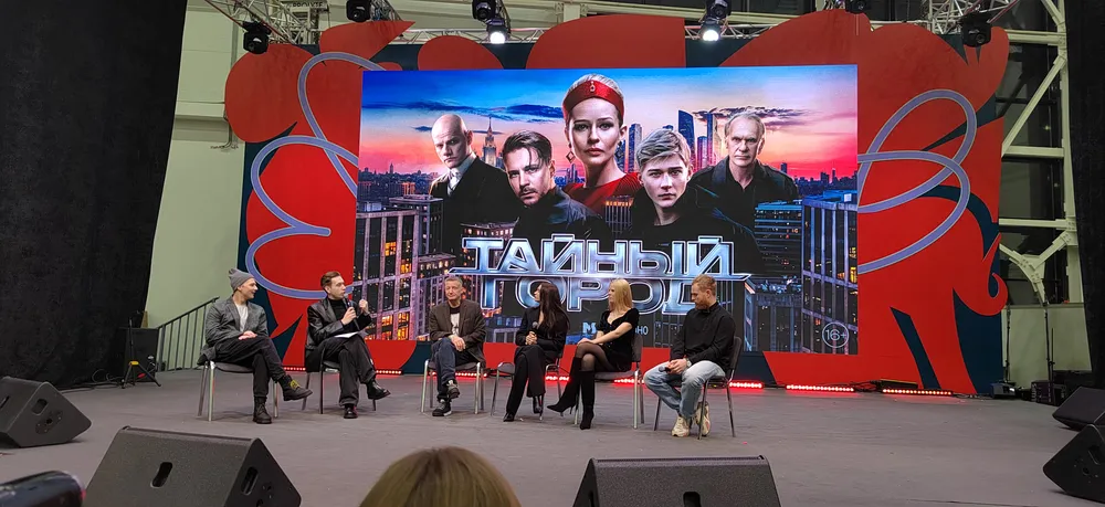 Обсуждение на сцене, группа людей, фон с изображением и названием, связано с публичным мероприятием.