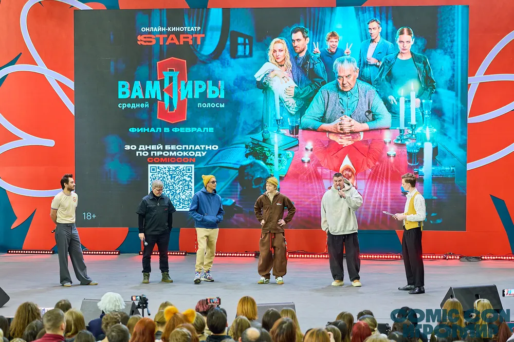 На изображении группа людей на сцене, возможно, в театральном представлении, с большим экраном на заднем плане с текстом и изображениями.