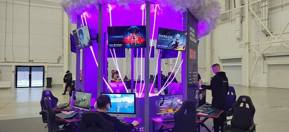 На картинке показан игровой стенд с несколькими мониторами и людьми, занимающимися играми в окружении яркой подсветки.