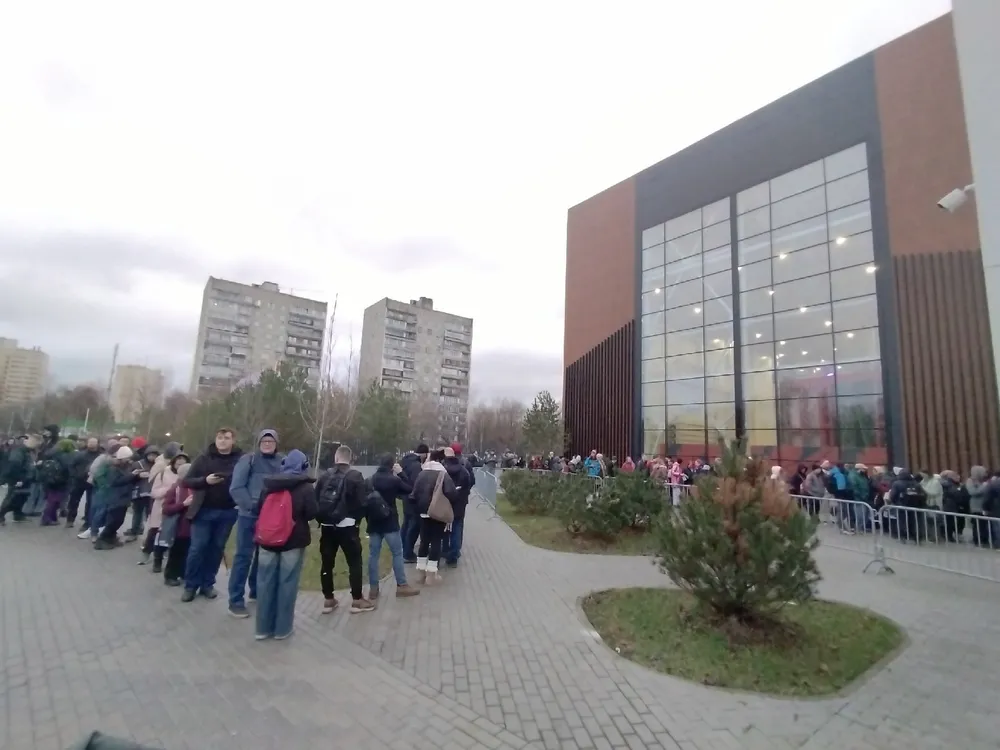 Очередь людей у здания, на заднем плане видны многоквартирные дома и кустарники.
