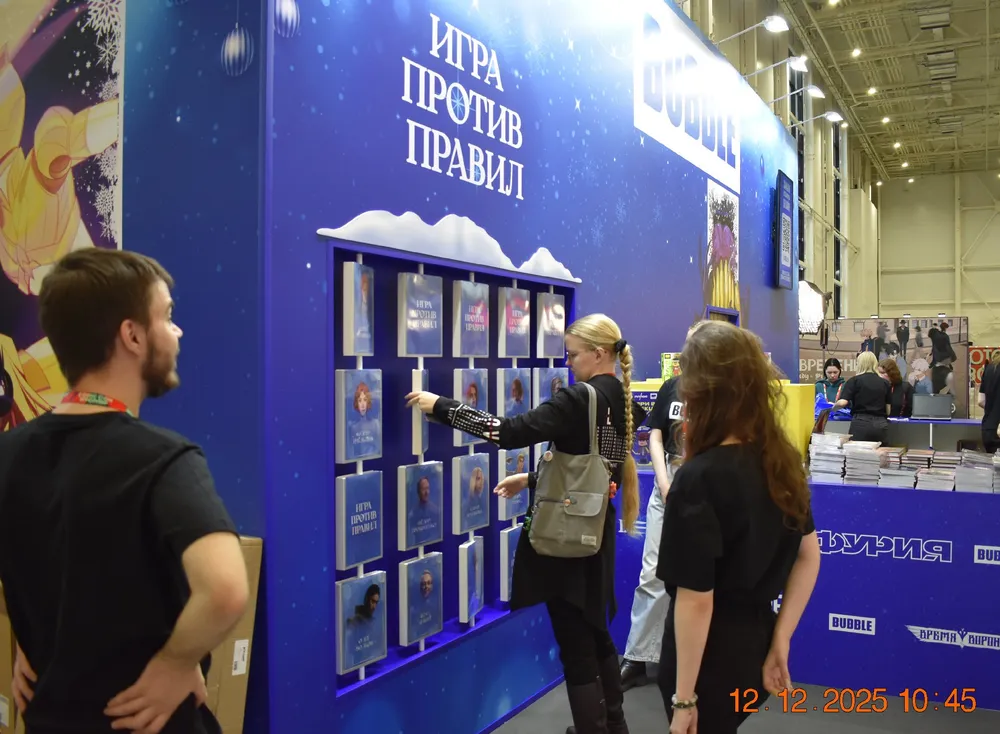 На изображении люди взаимодействуют с выставочной стенд, на котором расположены книги. Видно, что люди смотрят на стенд и обсуждают что-то.