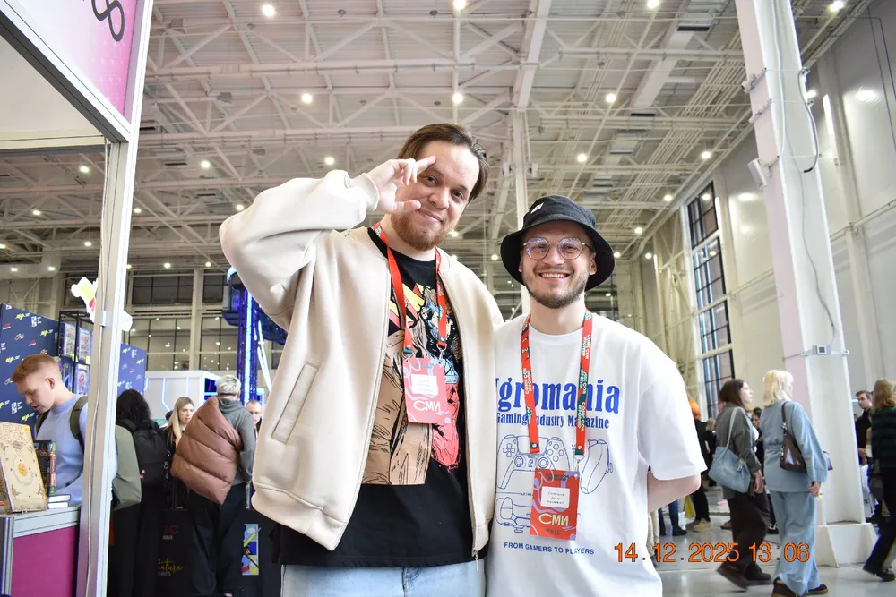 Два человека на мероприятии, один из них делает жест, другой улыбается. Окружение выставочного зала.