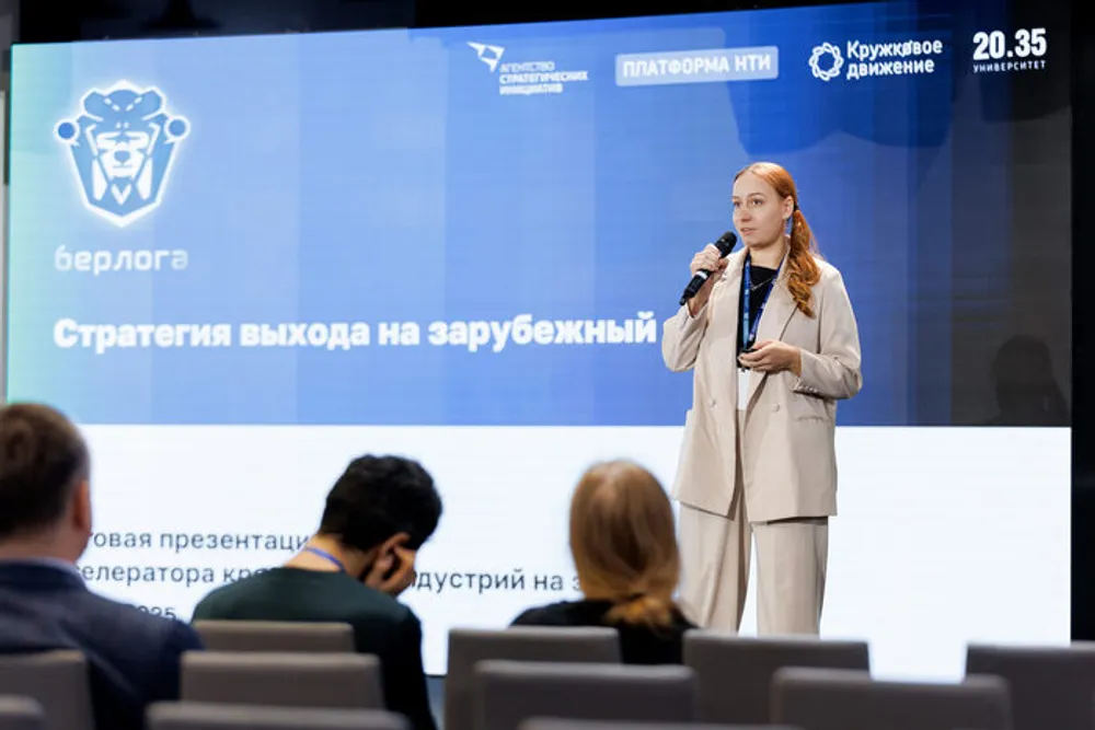 Изображение с женщиной, выступающей с речью на мероприятии. На заднем плане виден экран с текстом о стратегии выхода на зарубежный рынок.