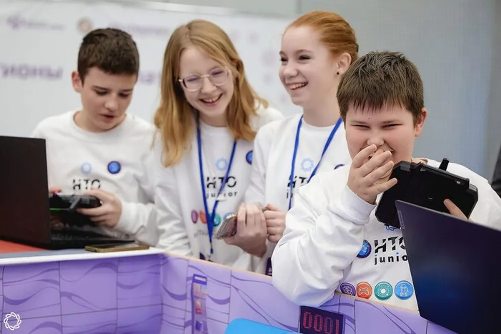 Группа подростков, улыбающихся и взаимодействующих с ноутбуками и игровыми устройствами на мероприятии.