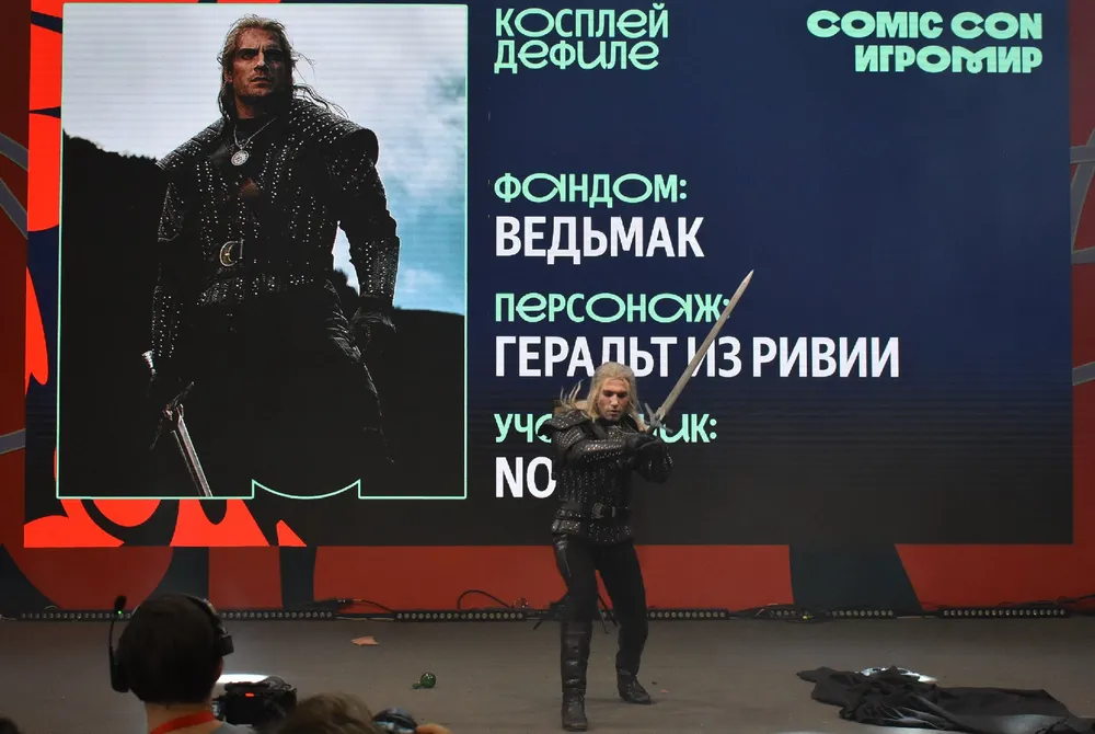 Изображение человека в костюме персонажа из фэнтези, с мечом, на фоне экрана с текстом о мероприятии. Упоминается персонаж \