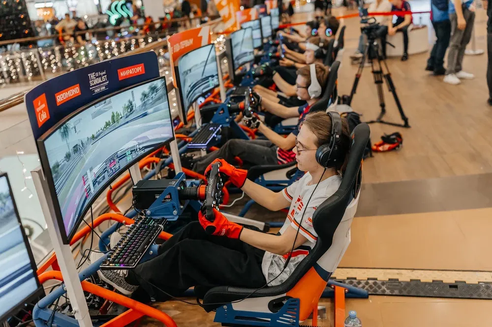 Группа людей, участвующих в соревнованиях по симуляторам вождения. Видны игровые установки и экраны.