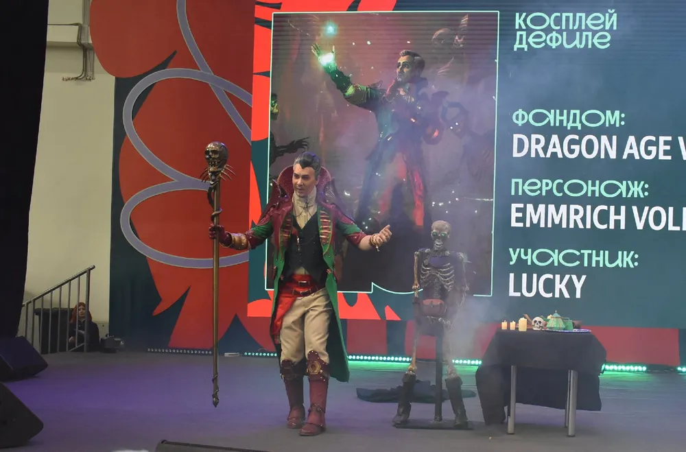 Косплей персонажа из игры Dragon Age, на сцене мужчина в костюме с черепом и магическим жезлом.