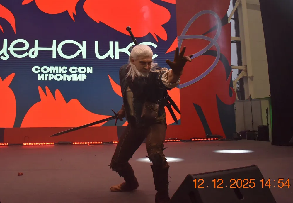 На изображении человек в костюме, позирующий на сцене с эффектной подсветкой, возможно, на мероприятии Comic Con.