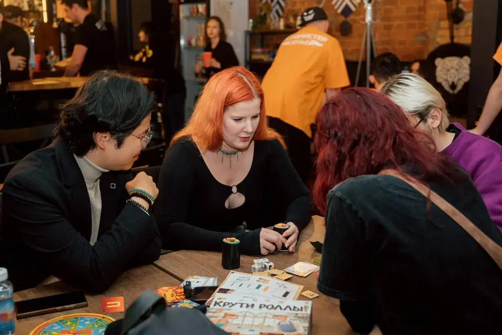 Группа людей за столом, играют в настольные игры. На столе видны игры и аксессуары для игры.
