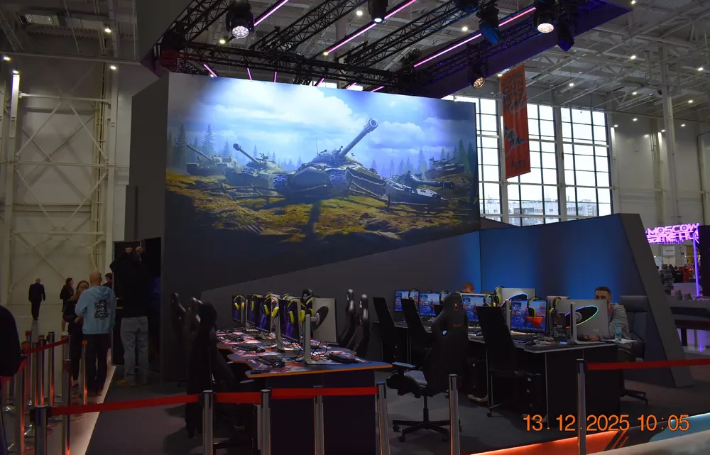 Изображение выставочного стенда с игровыми компьютерами и большим экраном, на котором показан пейзаж с военной техникой.