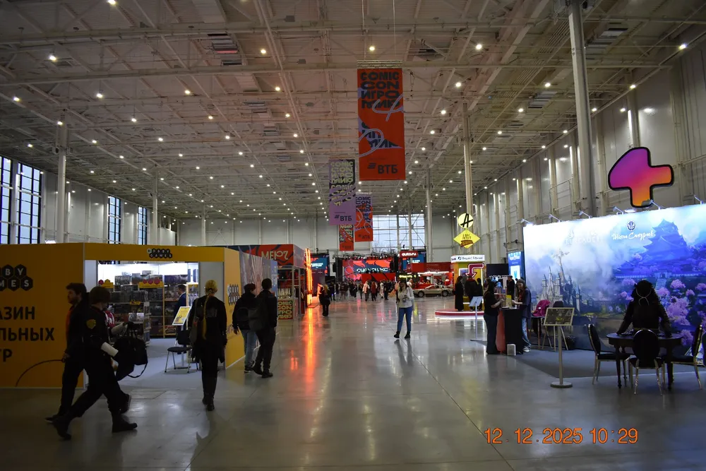 Изображение показывает интерьер выставочного зала с несколькими людьми, стендами и яркими вывесками.