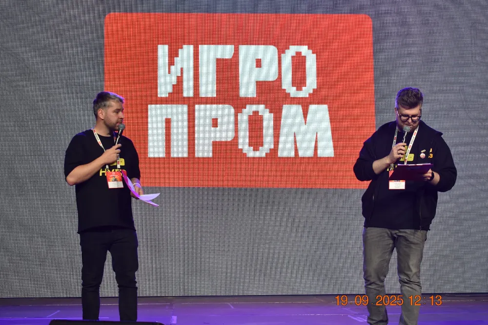 Два человека на сцене, один с микрофоном, другой читает с планшета. На заднем плане логотип \